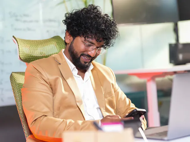 A professional at his desk, smiling at his phone beside a laptop